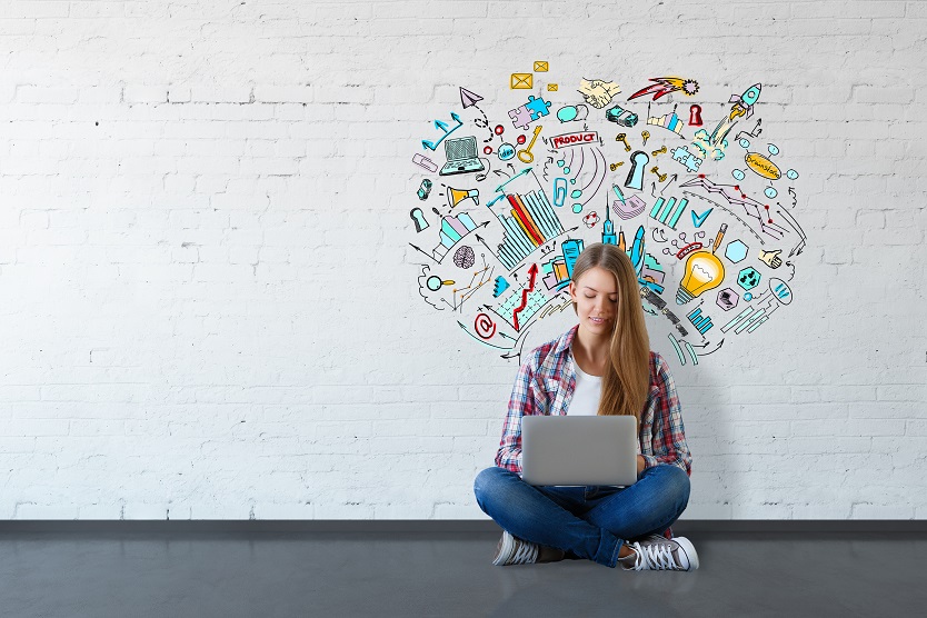 Junge Frau sitzt vor einer weißen Wand mit bunten Symbolen zum Thema Innovation und blickt auf ein Notebook.